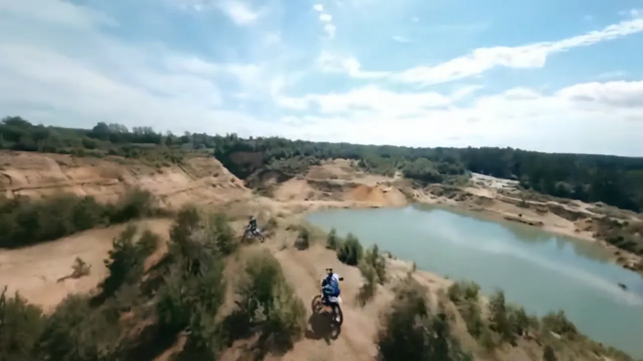 Two off-road riders on dirt bikes riding together along a trail beside a pond.